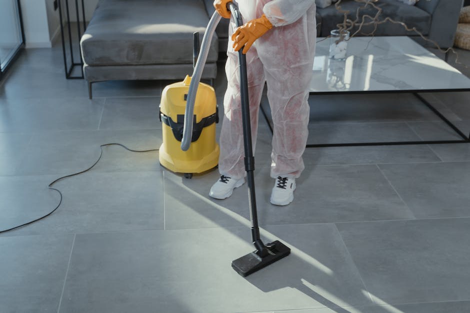 Two professional cleaning staff members from Petersham Carpet Cleaners stand indoors against a plain white wall, smiling and holding cleaning tools. The woman on the left, with dark hair styled in an