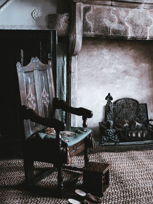 A room with a textured, patterned carpet covering the floor, featuring antique-style wooden furniture including a tall, intricately carved wooden chair with a upholstered seat, positioned next to a fi