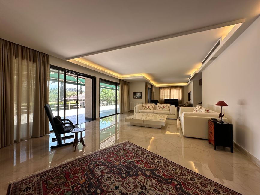 Bright, spacious living room with large bay windows featuring striped curtains, allowing natural light to illuminate the cream-colored walls and beige carpeted floor. The ceiling has a central ceiling